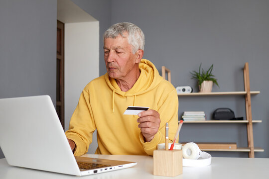 Serious senior man using laptop and holding credit card while sitting at table entering data during online shopping via pc computer making internet purchases