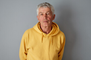 Serious gray haired Caucasian senior man wearing yellow hoodie standing isolated over gray background looking at camera with calm focused facial expression