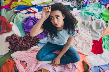 Above view full length photo of nice young girl arm touch head sit messy clutter clothes flat...