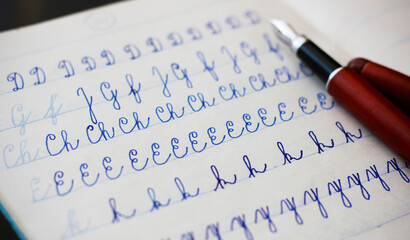 Close-up of uppercase and lowercase letters written in cursive with a fountain pen with blue ink in a lined notebook, a burgundy fountain pen in the background. Education and handwriting concept.