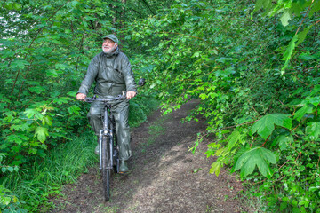 A smiling senior rides his bike through the rain-soaked forest in changeable weather. Wet branches hang low over the path. Well protected by rubber boots and rain gear, he enjoys the journey.