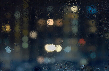 View through glass window with rain drops on blurred city lights at night