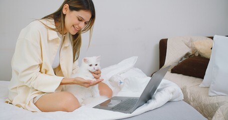 woman working on laptop online lying on bed in bedroom with her cat and petting cute pedigreed pet....