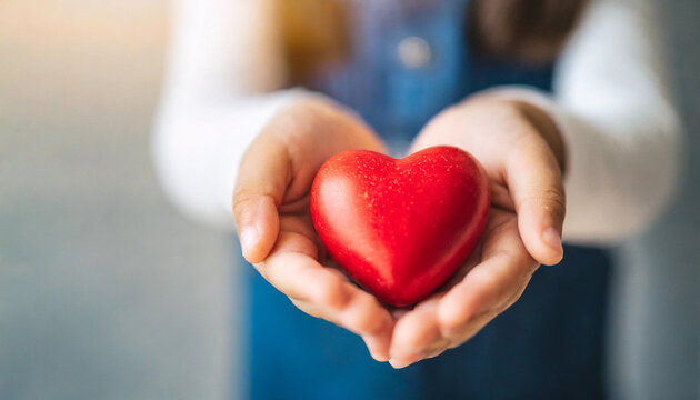 little girl's hand gently holds a bright red heart against a clean, empty, blurred wall background, symbolizing innocence, love, care, and hope in a pure and simplistic manner