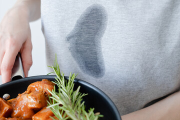 Splashing grease stain on gray clothes. An unrecognizable woman a frying pan with meat and herbs....