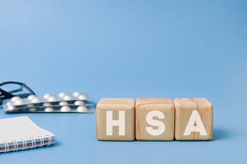 Wooden block with text HSA meaning Health Savings Account with glasses, notebook, pills behind on blue background
