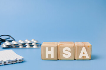 Wooden block with text HSA meaning Health Savings Account with glasses, notebook, pills behind on...
