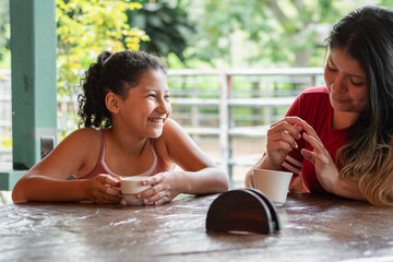 little brunette latina girl smiling, looking at her mother with pride while having a cup of coffee.