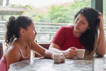 latina mother talking to her little daughter, very confused trying to explain to her while drinking coffee