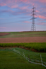 A large electricity pylon stands in a field, cables connected. The sky is colorful, with green...