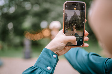 Riga, Latvia, - August 26, 2023 - A person takes a photo of an outdoor scene with a smartphone, capturing a statue and trees in the background.