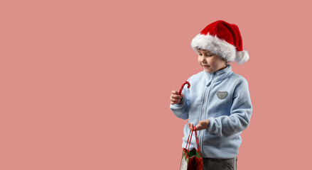 Christmas. A happy child in a Santa Claus hat holds a candy cane and a carrier bag. The background is pink. Copy space.