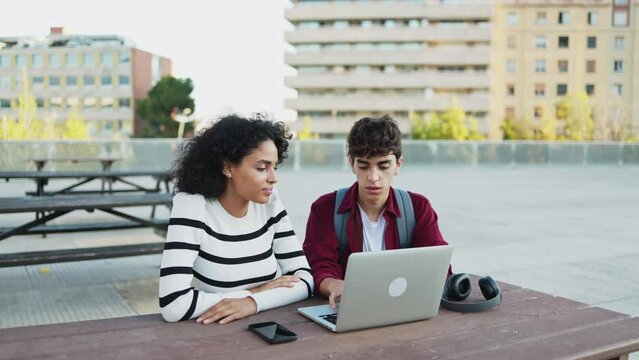 Young multi-ethnic students studying together on a laptop computer sitting on a bench on a university campus. Preparing for final exams. Concept of education and technology