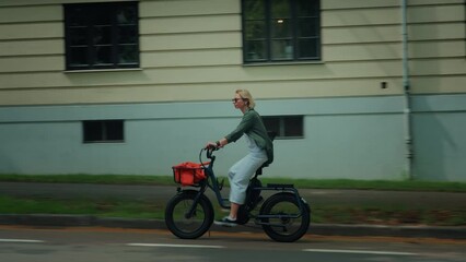 Handheld real life looped shot of woman ride cute electric city bike downhill on fast speed on busy street. cars pass by she rides on bike lane. Commuting and cycling in everyday life. Summer in city