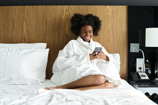 Portrait of young Black woman wearing bathrobe and using smartphone while relaxing on bed in hotel room, copy space
