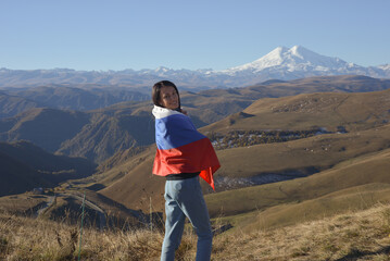 Fototapeta premium A young brunette woman stands against the backdrop of the snow-capped Mount Elbrus, looking at the camera, a Russian flag covers her shoulders. Tricolor against the backdrop of snow-capped Mount