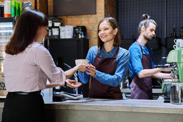 Woman customer of coffee shop near counter with cup of coffee talking to restaurant workers