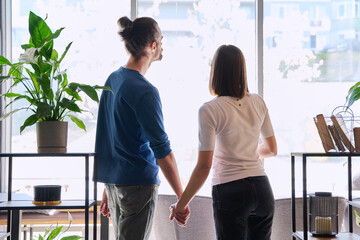 Back view, silhouette of young couple holding hands looking out window