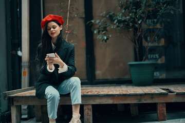 Stylish woman tourist in stylish clothes in a jacket and red beret sitting outside with phone in...