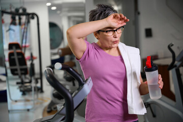 Fototapeta premium Mature tired woman wearing sportswear with bottle of water wiping her forehead