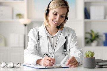 Smiling doctor in headphones having online consultation and filling documents at table indoors