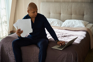 African american businessman with documents using laptop on bed in hotel room
