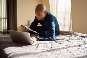 Focused african american male entrepreneur with documents using laptop on bed