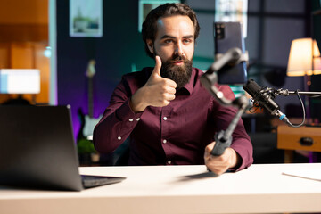 Content creator using cellphone on selfie stick to shoot video, doing thumbs up sign. Influencer making approval hand gesturing while filming vlog with mobile phone in cozy apartment