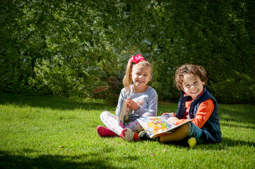 Irm&atilde;o sentados no gramado brincando e lendo livro, leitura infantil