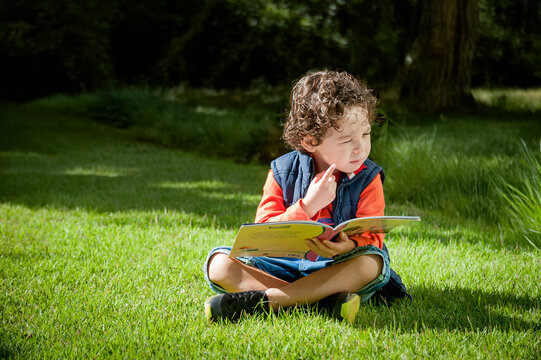 Menino sentado no gramado do jardim lendo, leitura infantil.