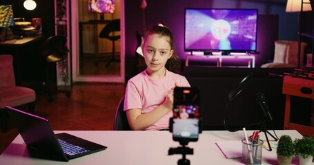 Zoom in shot of kid talking about school with children audience, filming content for social media with cellphone attached to tripod. Young girl recording video for internet channel with mobile phone