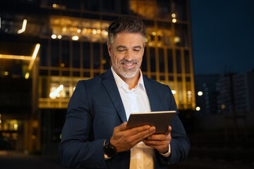 Closeup mature adult professional business man in formal suit working on pc computer. Smiling businessman ceo holding digital tablet using application at office building background in evening outdoors