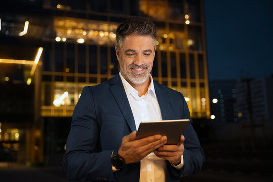 Closeup mature adult professional business man in formal suit working on pc computer. Smiling businessman ceo holding digital tablet using application at office building background in evening outdoors - Powered by Adobe