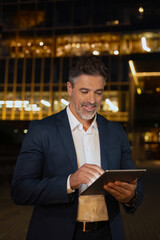 Successful mature adult professional business man in formal suit working on pc computer. Indian senior businessman ceo holding digital tablet using application outdoors at dark late time. Vertical