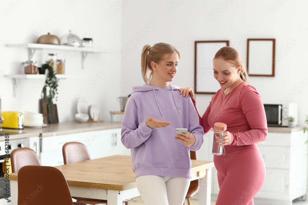 Sticker Sporty young women with smartphone drinking water in kitchen