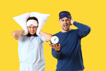 Displeased young couple with pillow and alarm clock on yellow background