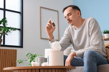 Ill young man with nasal drops sitting on sofa at home
