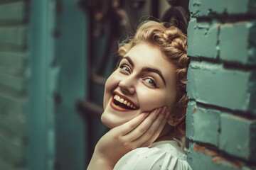 Fototapeta premium In this heartwarming image, a smiling young woman with curly blonde hair and bright eyes poses joyfully against a turquoise brick wall, showcasing genuine happiness and carefree spirit