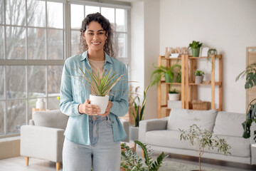 Beautiful young African-American woman holding plant in her hands in living room