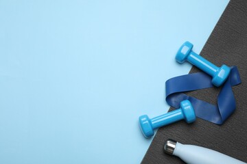 Two dumbbells, yoga mat, fitness elastic band and thermo bottle on light blue background, flat lay....