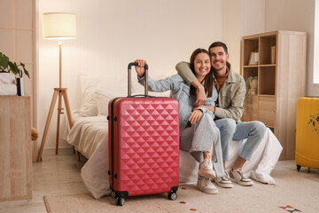 Young couple of tourists with suitcase sitting on bed in hotel room at night