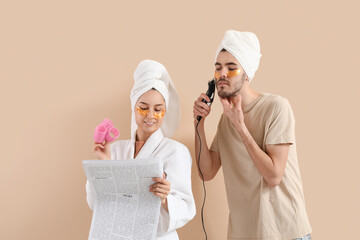 Young loving couple after shower with trimmer and hair curlers reading newspaper on beige background