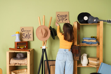 Young woman hanging cardboard with text GARAGE SALE on wall in room of unwanted stuff, back view