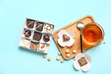 Delicious chocolate candies in box, wooden board, cup of tea, spoon and nuts on blue background