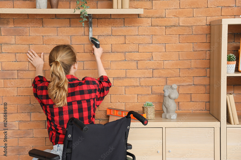 Poster Young woman in wheelchair drilling shelf at home, back view