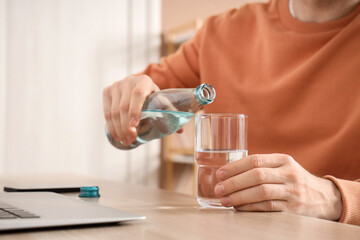 Young man pouring water into glass at table in office, closeup