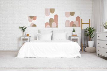 Interior of beautiful bedroom with comfy bed, bedside tables, chest of drawers and pictures on white brick wall