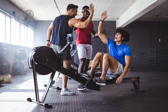 At gym, three young, fit, diverse men are high-fiving near rowing machine