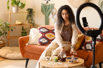Young African-American witch with magic attributes recording video at home