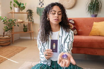 Young African-American witch with tarot card and stone at home